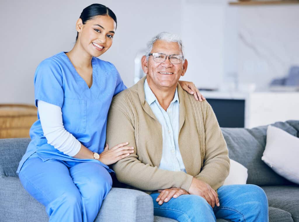 A smiling female caregiver in blue scrubs sitting beside an elderly man with glasses, who is wearing a beige cardigan, in a cozy home setting. Both are looking at the camera with warmth and trust. home care pittsburgh pa