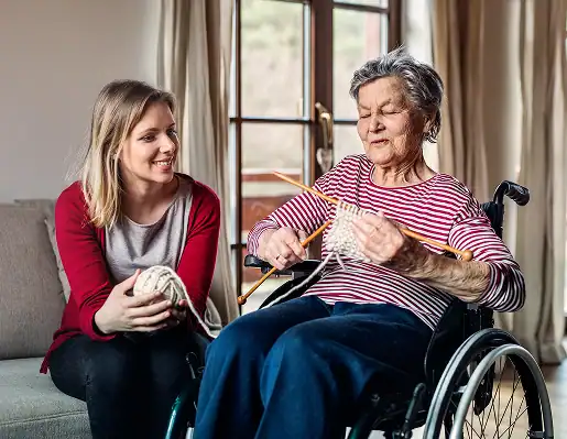 Elderly woman in wheelchair knitting, with caregiver. Elderly care services at home.