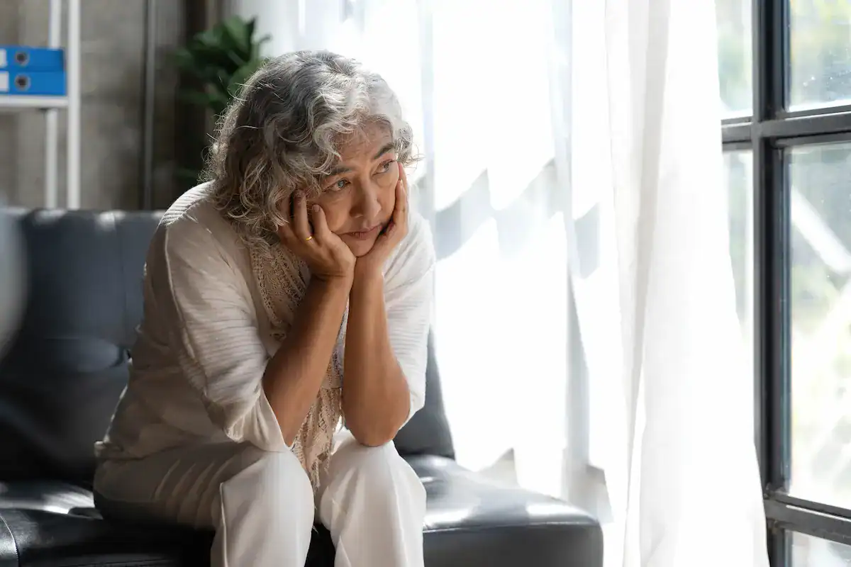Elderly woman sitting on couch, looking sad, touching her face